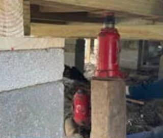 Red hydraulic bottle jack on a wood post pushing up a floor beam beside concrete block piers